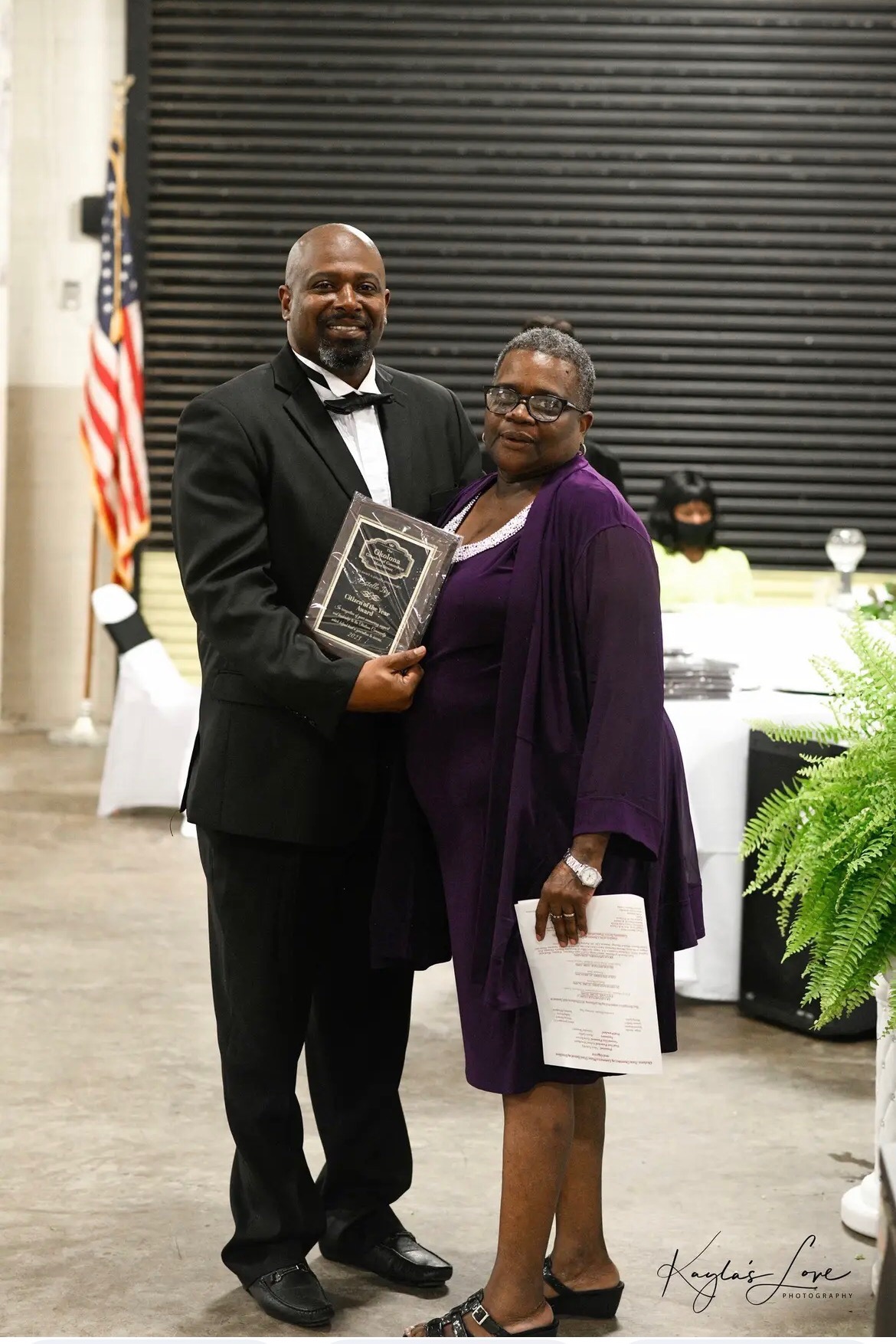 A man and woman stand side by side. The man holds a plaque which notes the woman as Citizen of the Year. Photograph by Kayla's Love Photography. 